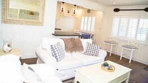 A living room with a white sofa, checkered pillows, a coffee table with decorative items, a mirror with an ornate frame, and a kitchen area with pendant lights and a clock. - 4980 Sandyland Road #216 2 Bedroom Condo by RedAwning (Carpinteria)