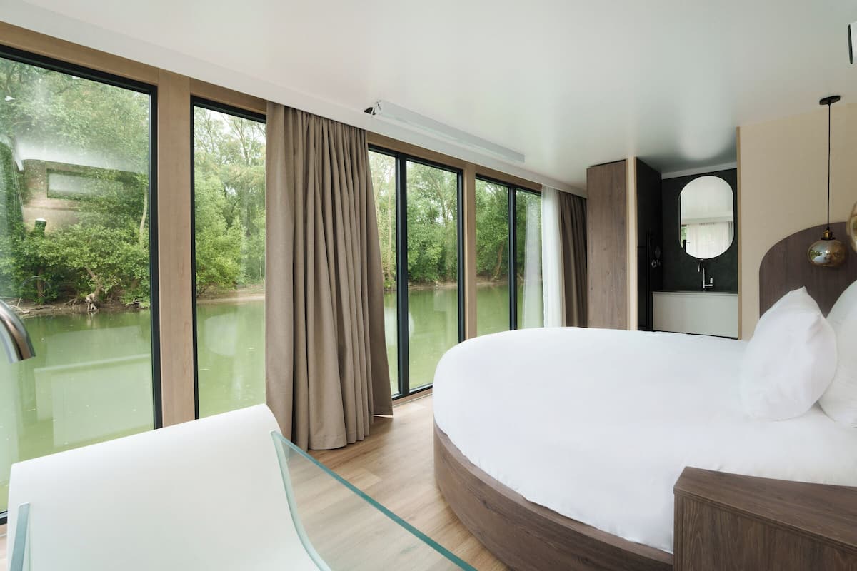 A modern hotel room with a large bed, a glass table, and a view of a river.