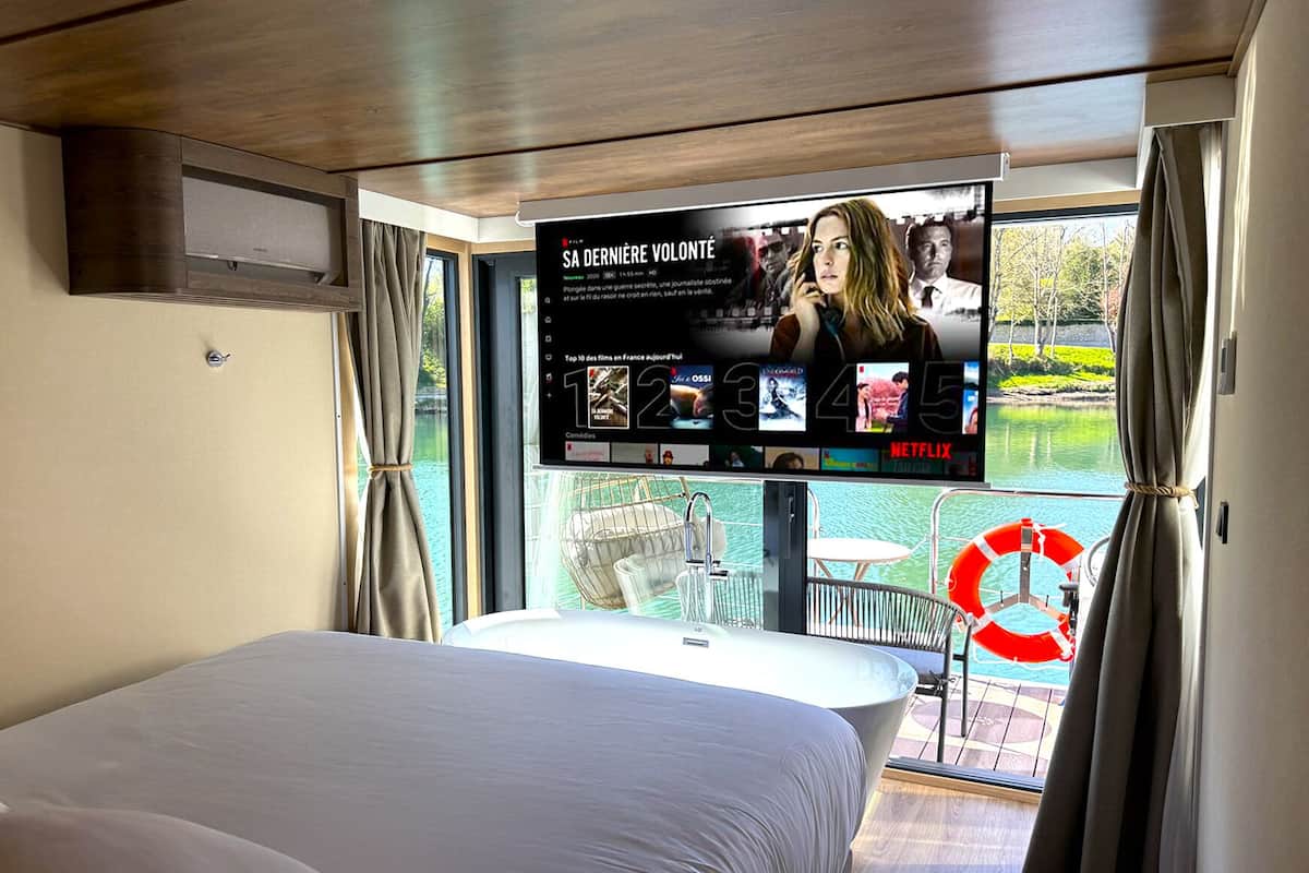 A modern hotel room with a large bed, a TV mounted on the wall displaying a movie, and a view of a lake through the window.