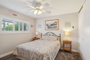 A bedroom with a bed, two bedside tables with lamps, a ceiling fan, a window with blinds, and a framed picture on the wall.