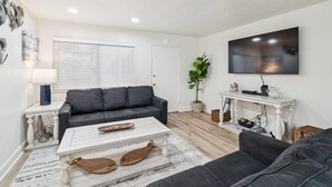Una sala de estar con un sofá gris, una mesita de centro blanca, un televisor en la pared y una planta en maceta.