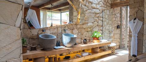 Une salle de bain aux murs de pierre, dotée de deux grands lavabos en pierre, d’un meuble-lavabo en bois et d’un miroir qui reflète la pièce.