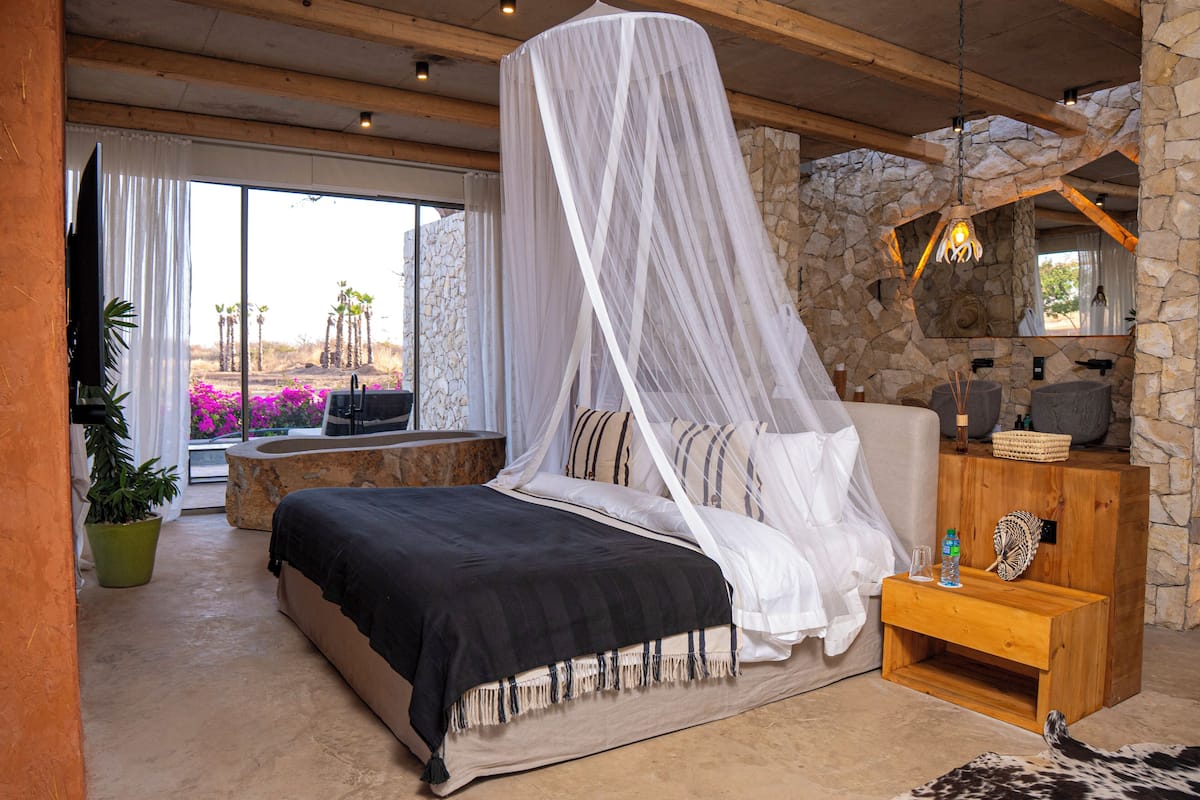 Quarto com cama, parede de pedra, mesa de cabeceira de madeira, planta em vaso e vista para palmeiras e flores.