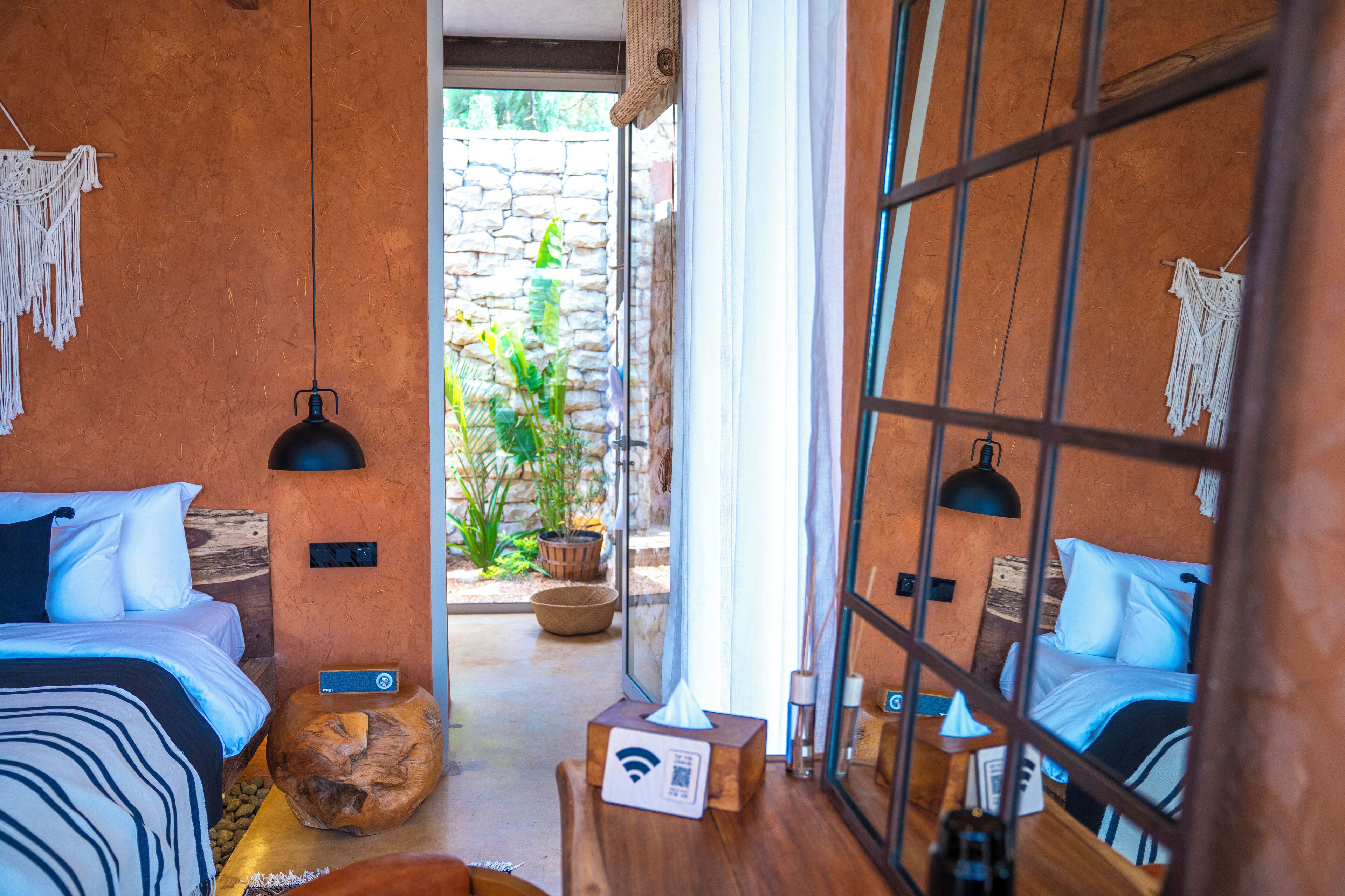 Una habitación de hotel moderna con dos camas, una pared de piedra, piso de madera, una mesita con una estación de carga inalámbrica y vistas a un jardín a través de un ventanal.