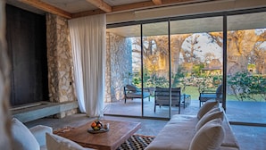 A modern living room with a stone wall, a sofa, a coffee table with fruit, and a view of an outdoor patio with furniture. - Casa del Toro Luxury Ranch (Nguekhokhe)