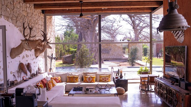 A spacious living area with a large window, a white sofa, a coffee table, and a stone wall with mounted animal heads. - Casa del Toro Luxury Ranch (Nguekhokhe)