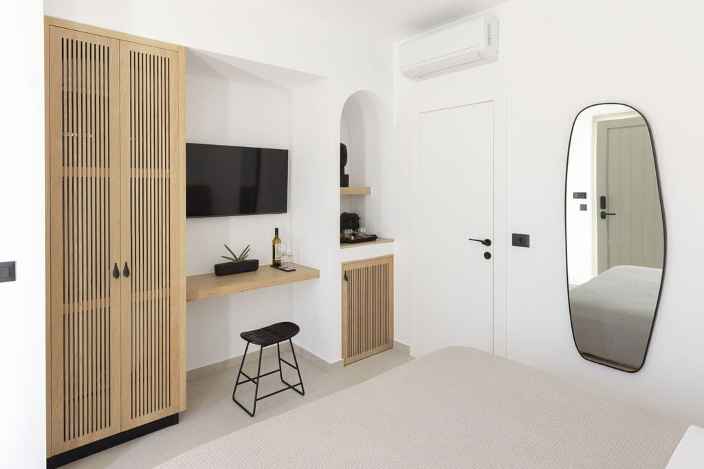 A modern bedroom with a wooden wardrobe, a mounted TV, a small desk with a plant, and a large mirror.