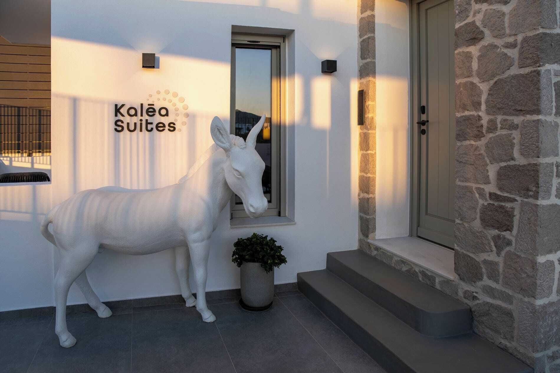 A building with a sign reading 'Kaléa Suites,' a white donkey sculpture, a potted plant, and a stone wall.