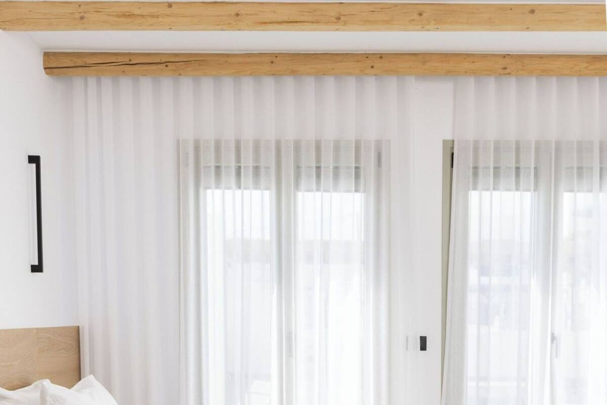 Ein modernes Schlafzimmer mit einem Holz-Kopfteil, einem großen Bett mit weißer und beiger Bettwäsche und transparenten weißen Vorhängen.