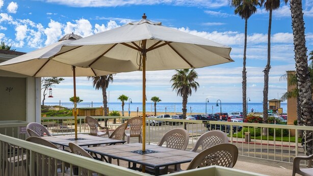 A balcony with a table and chairs, overlooking a beach with palm trees and parked cars.