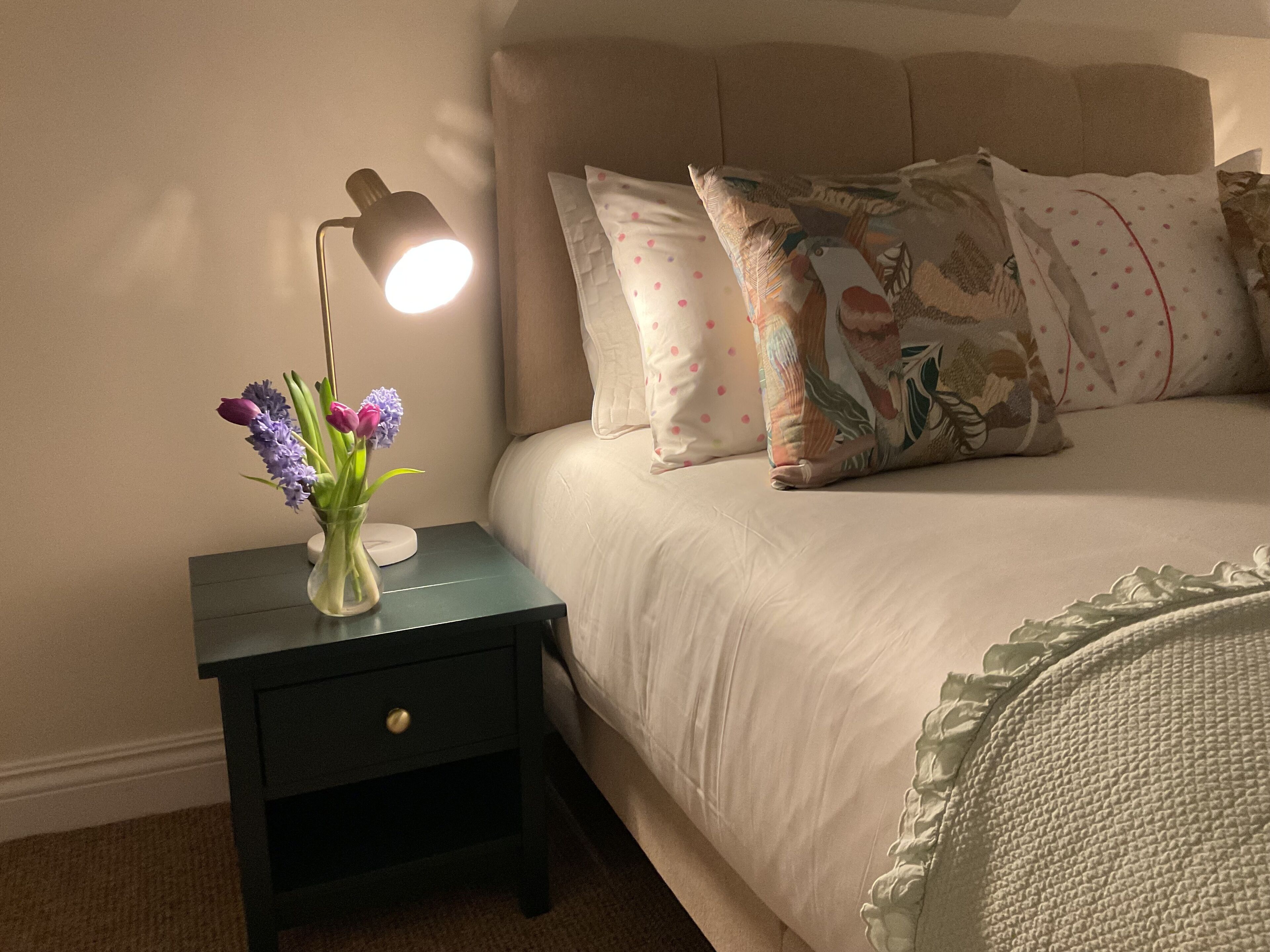 Une chambre à coucher avec un lit, des oreillers, une table de chevet, une lampe et un vase rempli de fleurs.