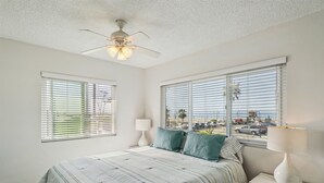 A bedroom with a bed, two bedside lamps, a ceiling fan, and large windows with blinds.