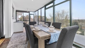 Een eetruimte met een lange tafel gedekt voor vier personen, grote ramen en uitzicht op de omgeving.