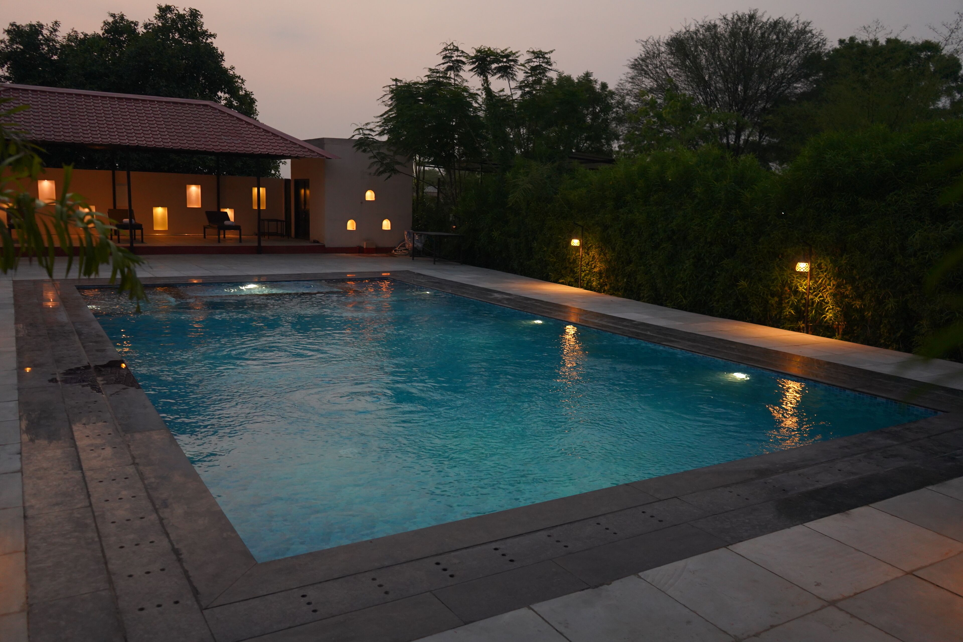 Una piscina con borde de baldosas, rodeada por un seto bajo y un edificio con tejado de azulejos.
