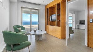 Un salon moderne avec une table ronde, deux fauteuils verts, un meuble de rangement en bois avec un téléviseur, et une vue sur la mer par une porte vitrée.