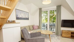 Ett modernt vardagsrum med en grå fåtölj, ett träbord, en soffa och en tv. Det finns en trappa till vänster och ett fönster med gardiner.