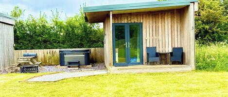 A modern wooden cabin with a covered patio, a barbecue grill, and two chairs.
