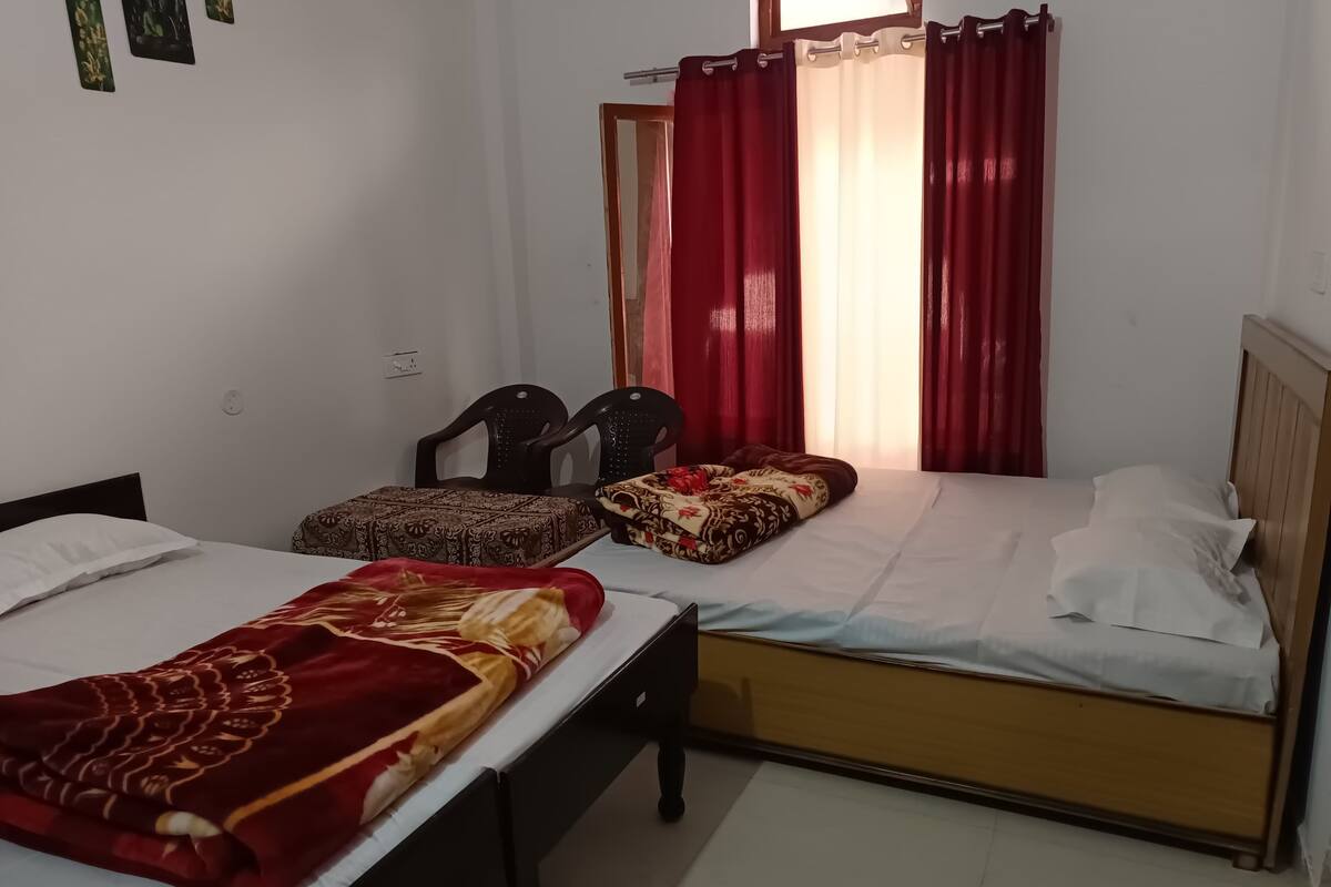 Quarto com duas camas, uma com manta vermelha e outra com roupa de cama branca. Há duas cadeiras e uma pequena mesa.