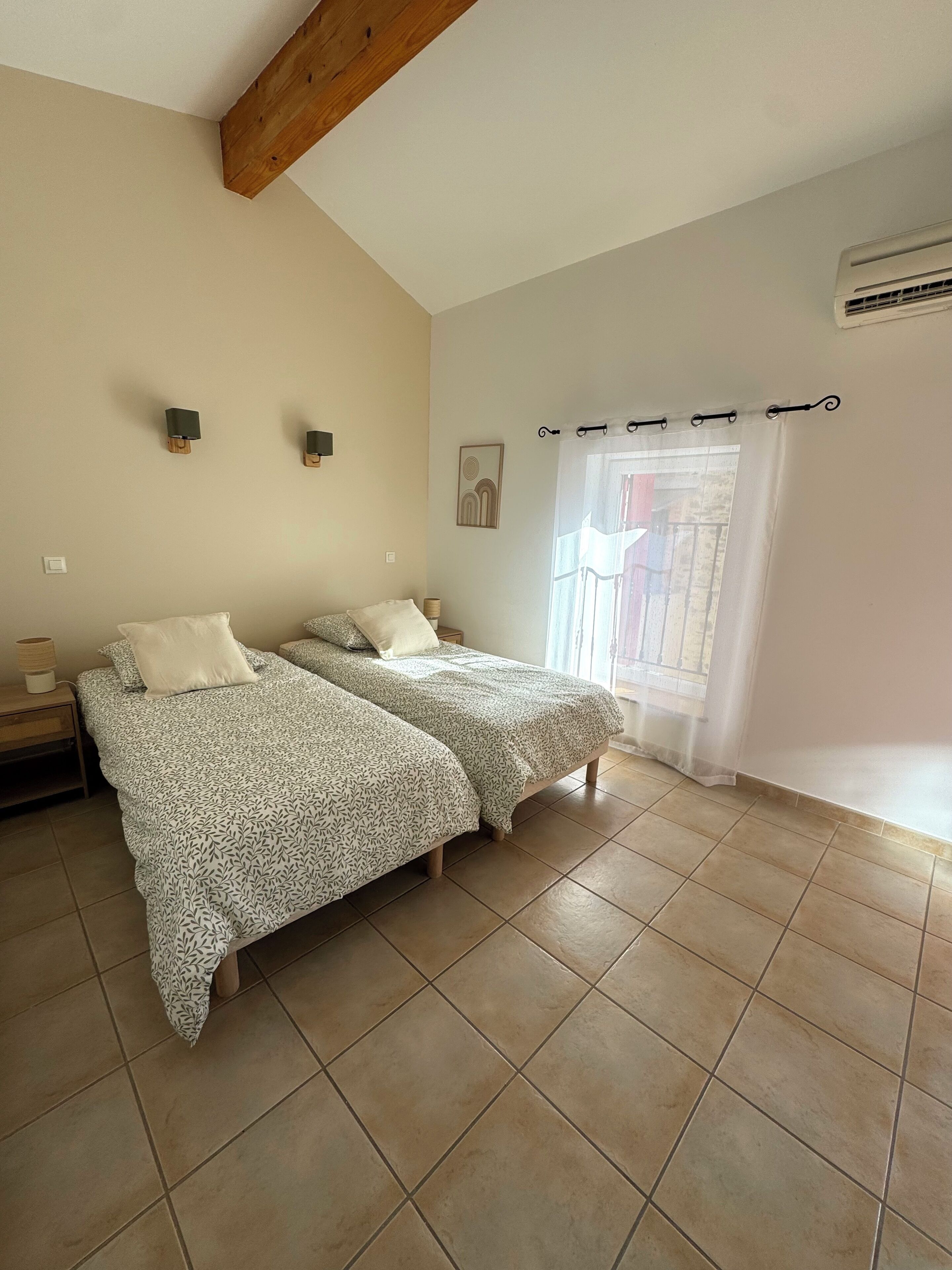 A bedroom with two beds, a wooden ceiling beam, a wall-mounted air conditioner, and a window with a sheer curtain.