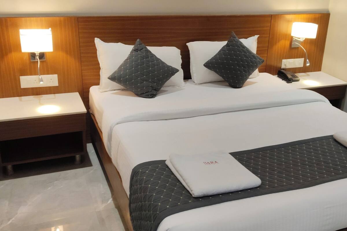 A neatly made bed with white linens, two dark pillows, and a folded towel on the bed. There are two bedside tables with lamps, a wooden headboard, and a nightstand with a phone.
