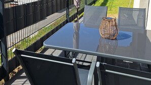 Eine Terrasse im Freien mit einem Glastisch, Metallstühlen und einer Lampenschirm aus Korbgeflecht.