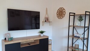 Ein modernes Wohnzimmer mit einer Holzeinbauwand, einem Flachbildfernseher, einem dekorativen Wandbehang aus Makramee und einem schwarzen Metallregal mit diversen Gegenständen.