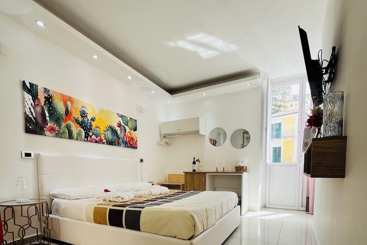 Ein modernes Schlafzimmer mit einem großen Bett, einem Bild mit Kakteen, einer Klimaanlage und Blick ins Freie.
