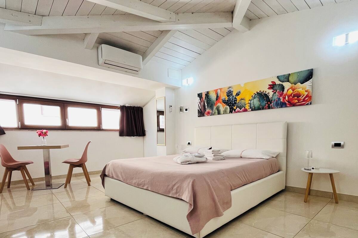 Ein modernes Schlafzimmer mit einem großen Bett, einem kleinen Tisch, zwei Stühlen, einem Fenster und einem Wandbild mit Kakteenmotiven.