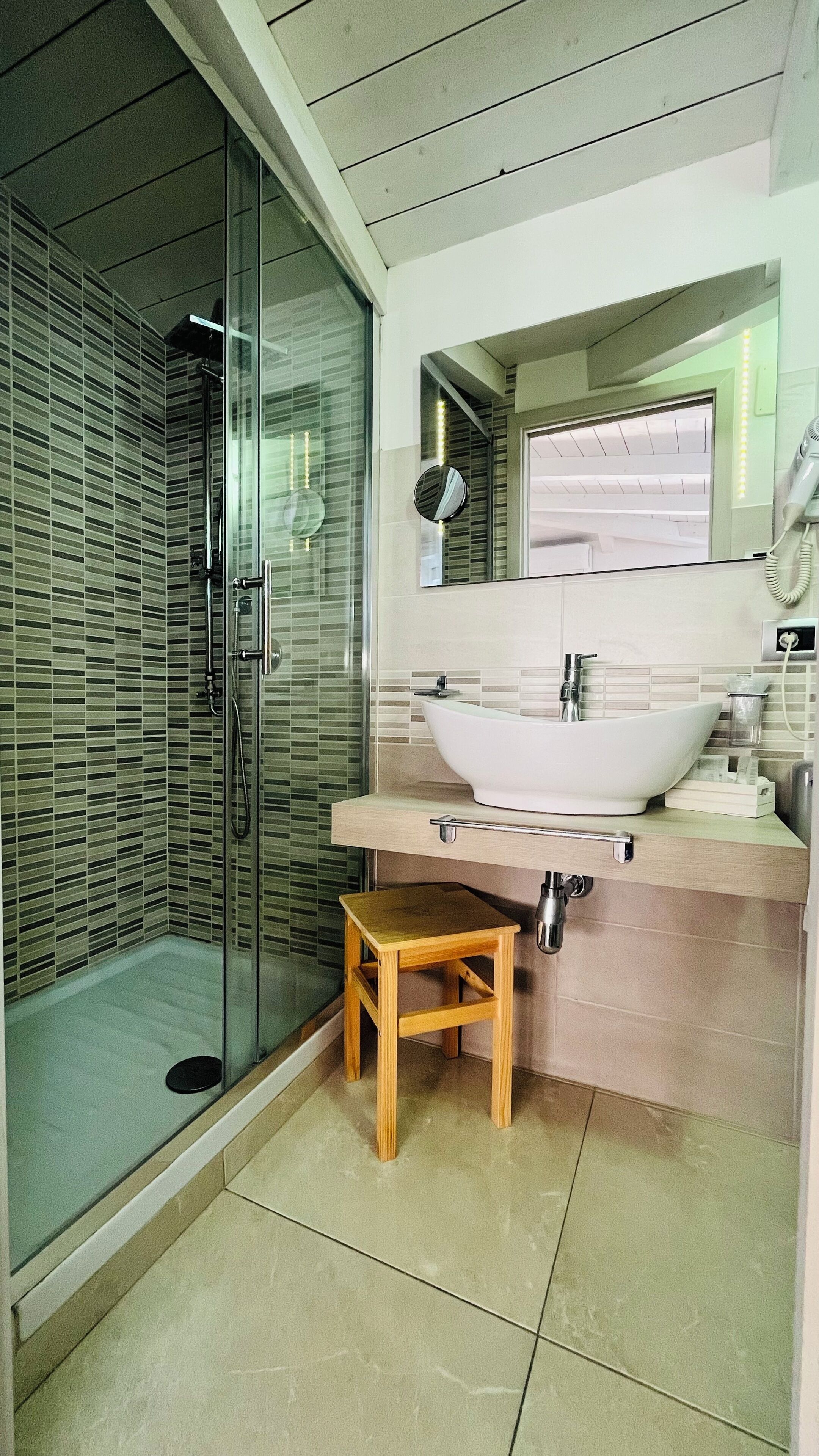 A modern bathroom with a glass-enclosed shower, a wooden stool, a white sink, and a mirror above the sink.
