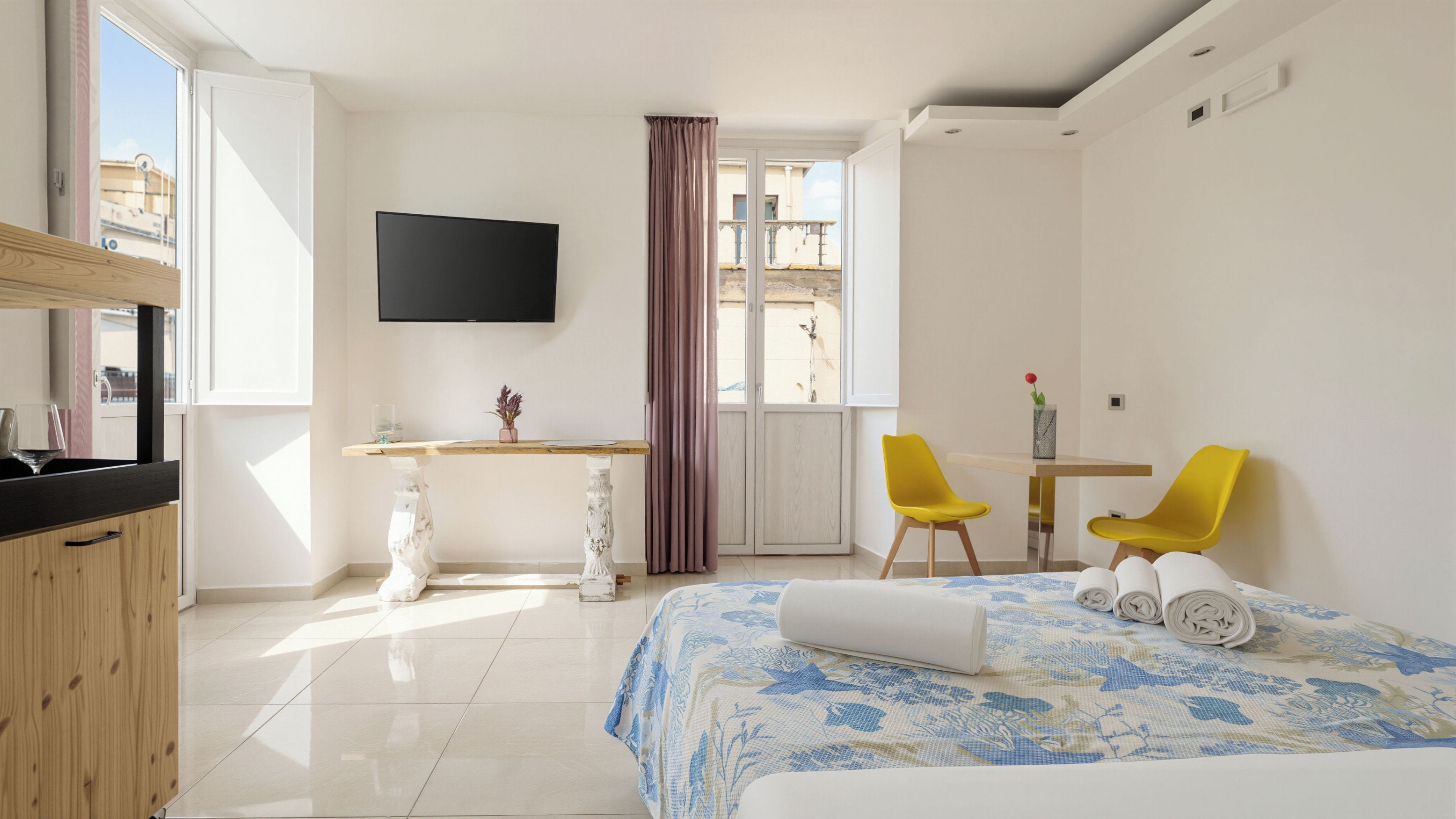 Une chambre d’hôtel avec un lit, un bureau, deux chaises, une télévision et une fenêtre avec des rideaux.