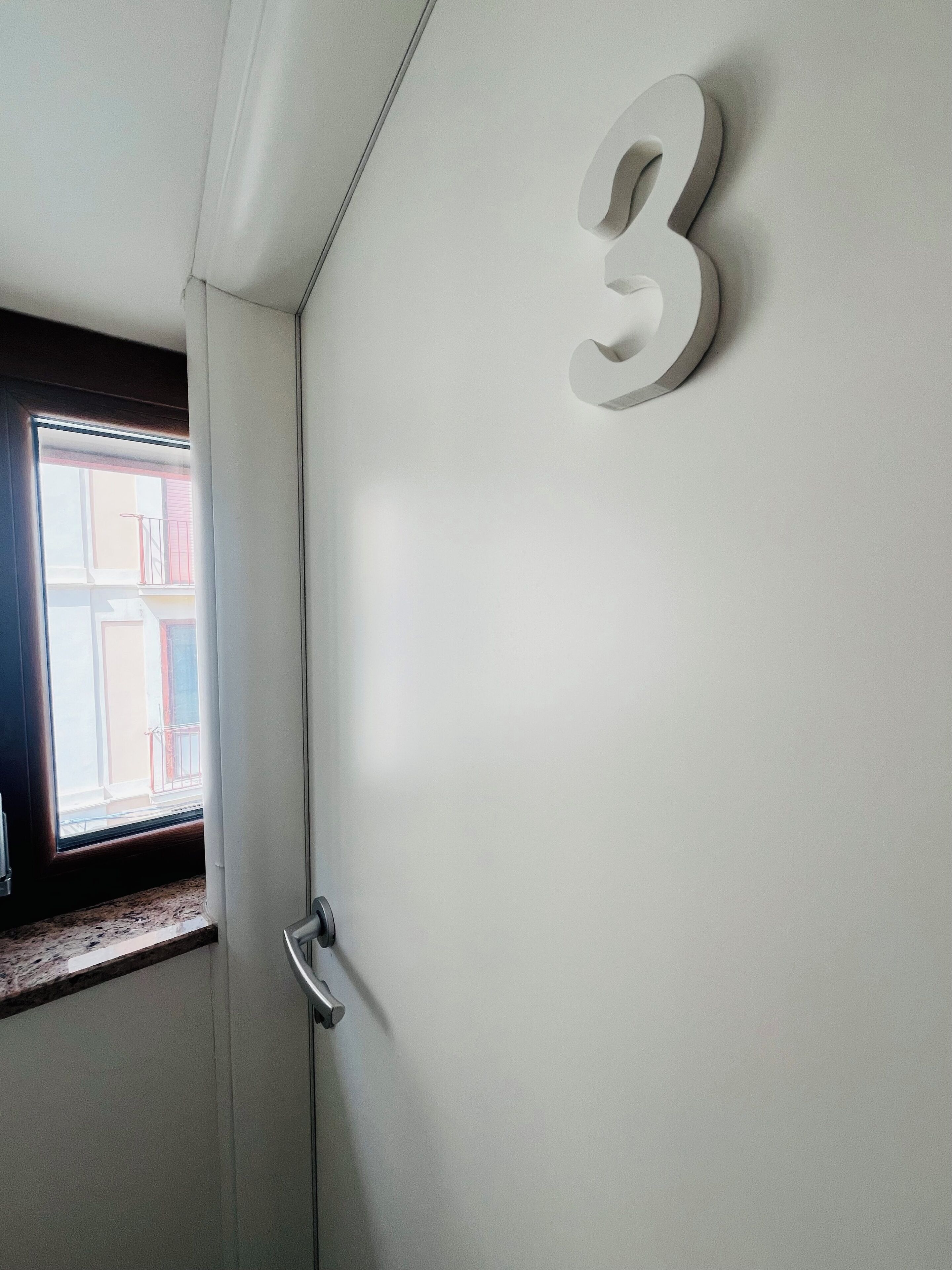 Une porte avec une plaque portant le numéro « 3 », une fenêtre donnant sur un balcon et une poignée de porte.