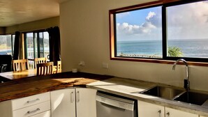 A kitchen with a view of the ocean, wooden cabinets, a dishwasher, and a dining area with chairs. - 90 Mile Paradise  (Kaitaia)