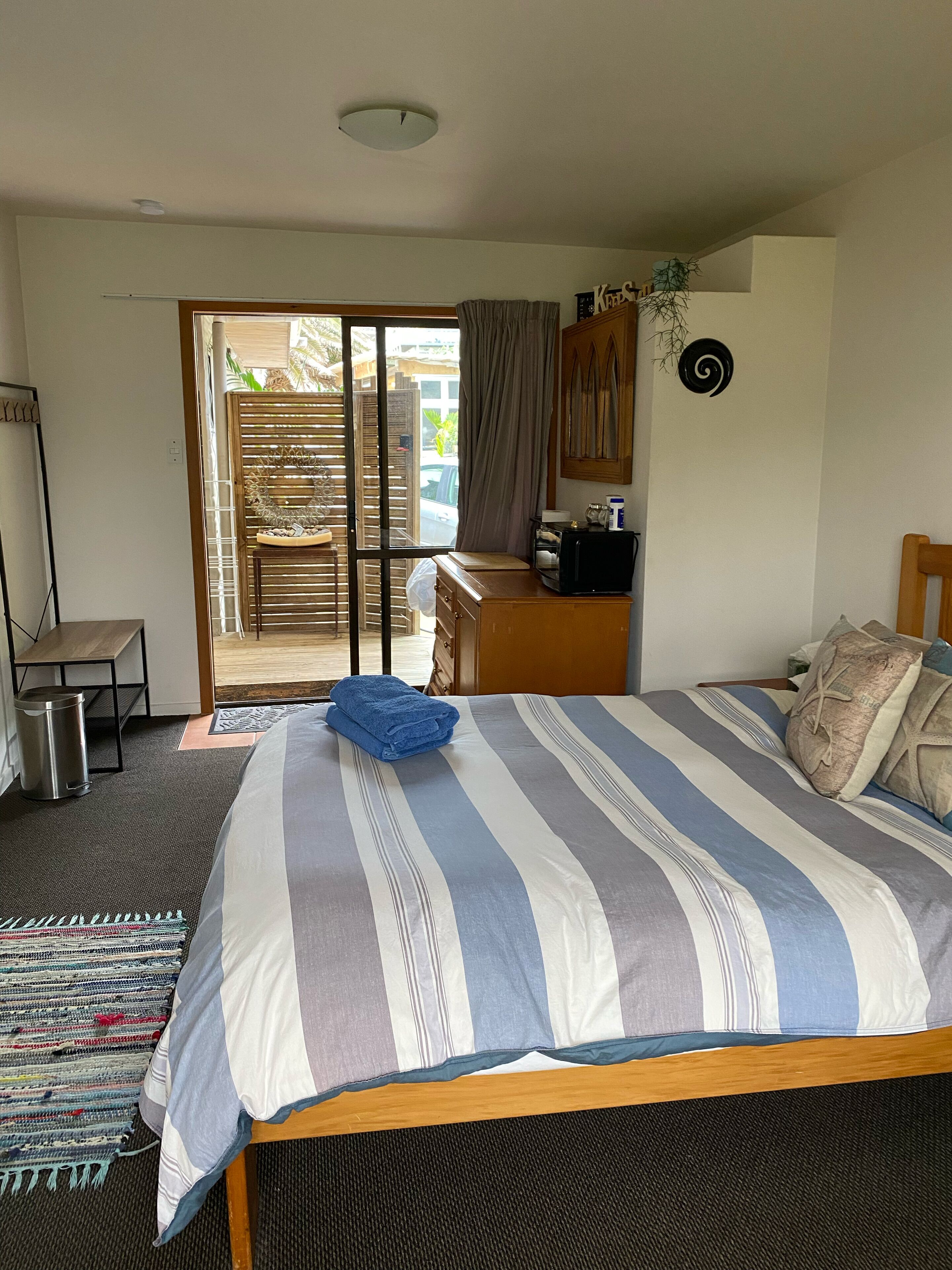 Une chambre à coucher comprenant un lit, un bureau, une chaise, une poubelle et donnant sur l’extérieur.