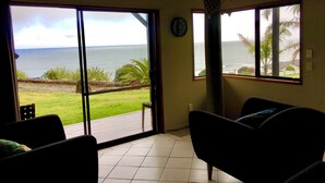 A room with a large sliding glass door leading to a balcony with a view of the ocean. There is a ceiling fan, a clock on the wall, and a dark-colored sofa. - 90 Mile Paradise  (Kaitaia)