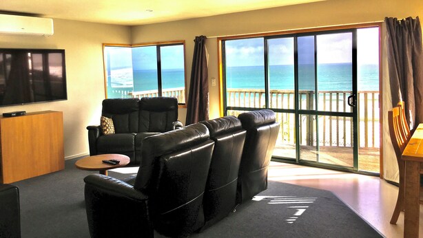A living room with a large TV, a sofa, and a view of the ocean through large windows. - 90 Mile Paradise  (Kaitaia)