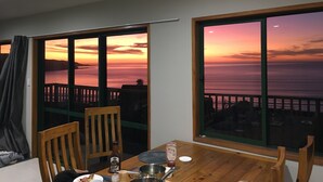 A dining area with a table set for two, a bottle of wine, and a view of the sunset over the ocean. - 90 Mile Paradise  (Kaitaia)
