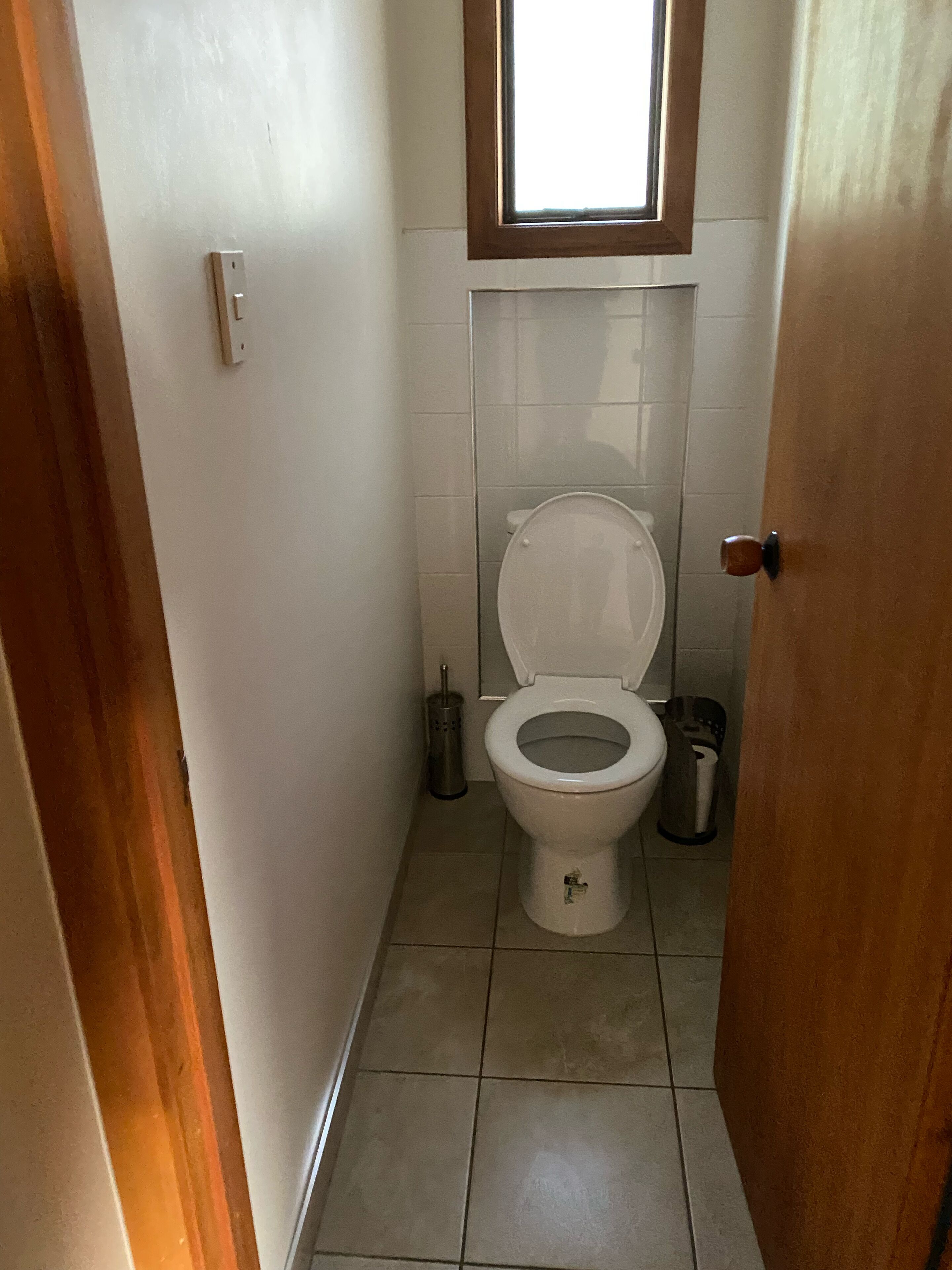 A small, narrow bathroom with a toilet, a window above the toilet, and a door on the right.