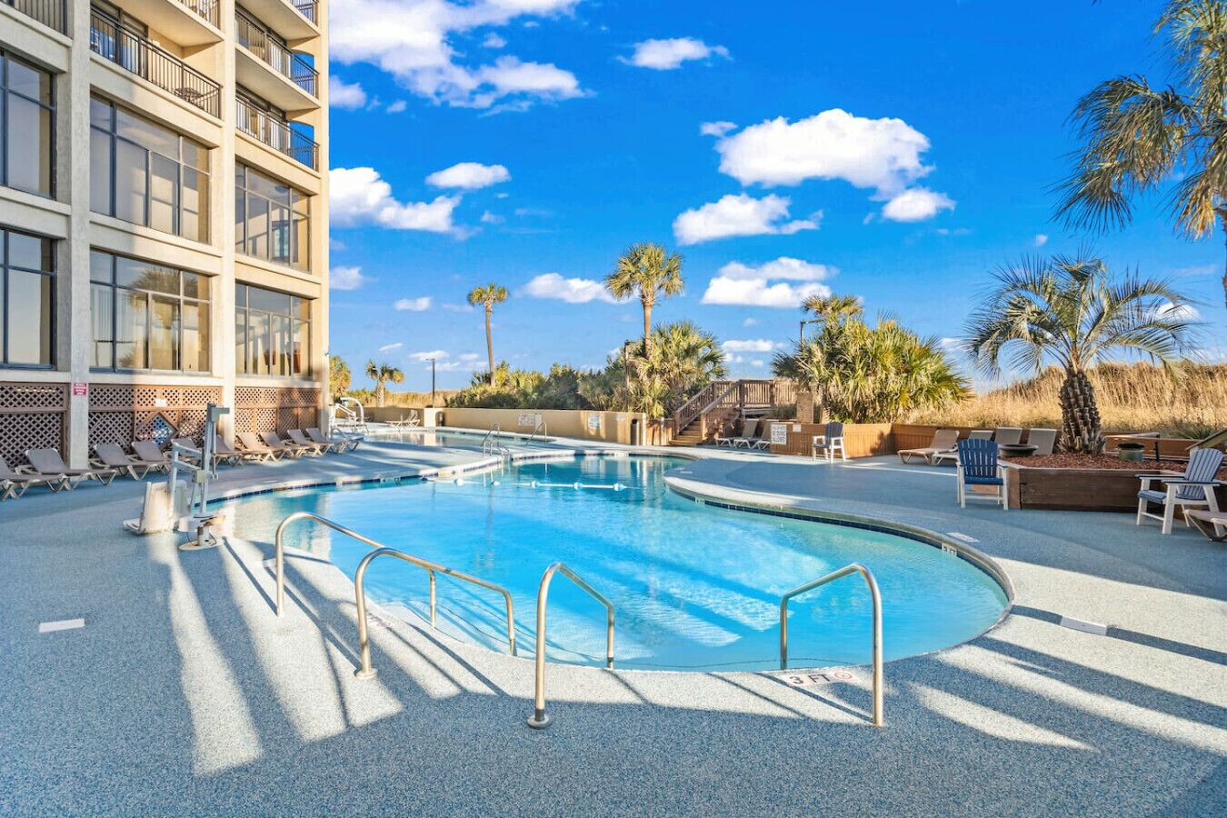 A swimming pool with sun loungers and palm trees, surrounded by a building with balconies.