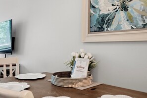 A dining area with a table set for two, a wicker basket with a guest book, and a framed abstract painting on the wall.