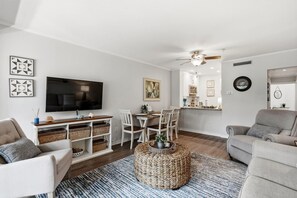 A modern living room with a flat-screen TV, a dining area with a table and chairs, a kitchen area with a microwave, and a ceiling fan.