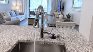 A modern kitchen with a granite countertop, stainless steel faucet, and a sink. There is a dining area with a table and chairs in the background. - Oceanfront Resort Condo with Relaxing Pool Hot Tub (Myrtle Beach)