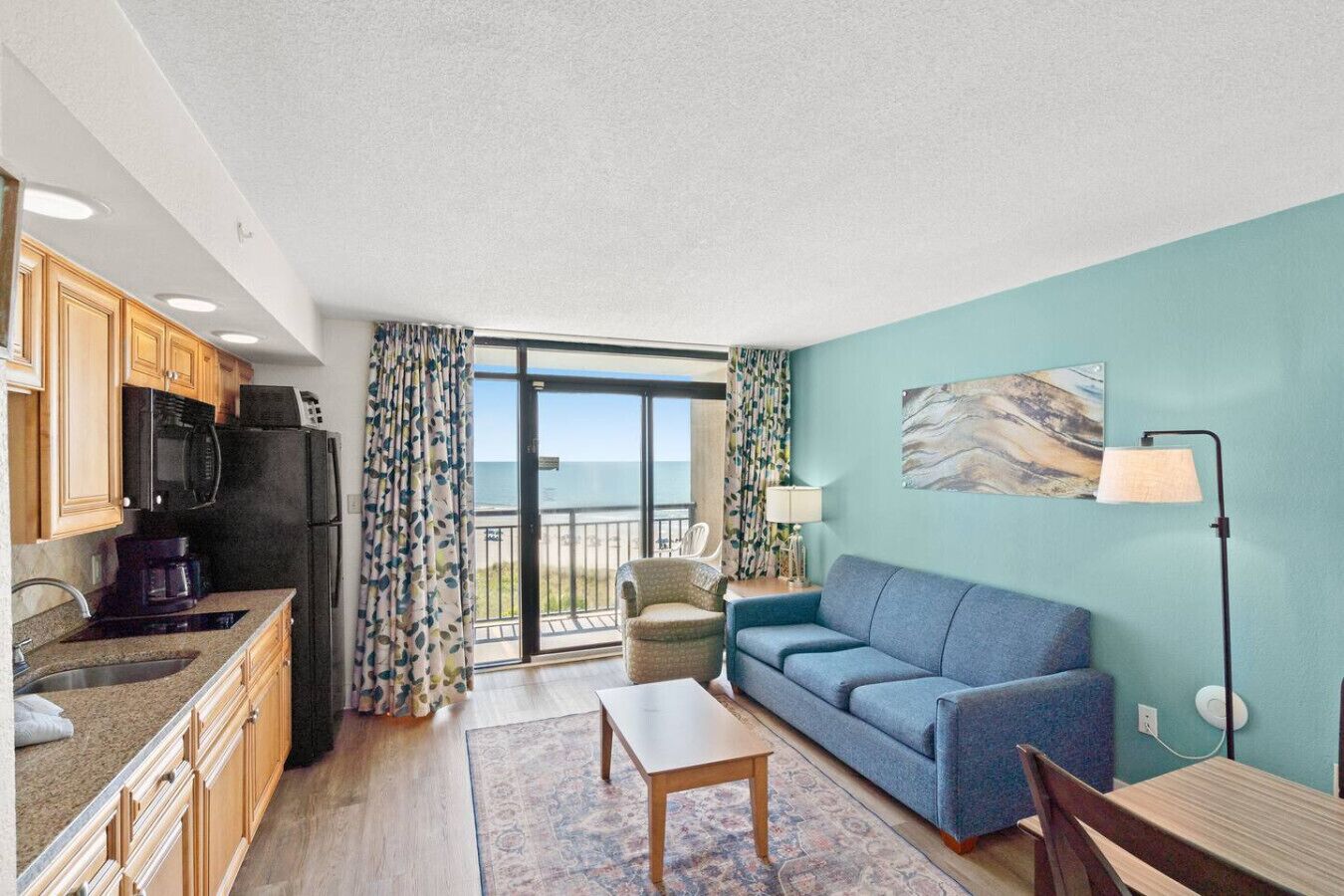 Un espacio habitable pequeño con kitchenette, un sofá, una mesa de comedor y vista al océano por un ventanal corredizo.