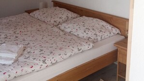 Un dormitorio con estructura de cama de madera, colchón blanco con motivos florales, mesita de noche de madera y un cuadro de flores en la pared.
