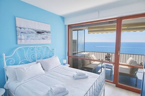 A bedroom with a bed, white pillows, a nightstand, a painting of the sea, and a balcony with a table and chairs overlooking the ocean. - I Gabbiani sul Mare (Cervo)