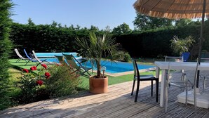 En træterrasse med swimmingpool, liggestole, et bord og en stråtækt parasol.