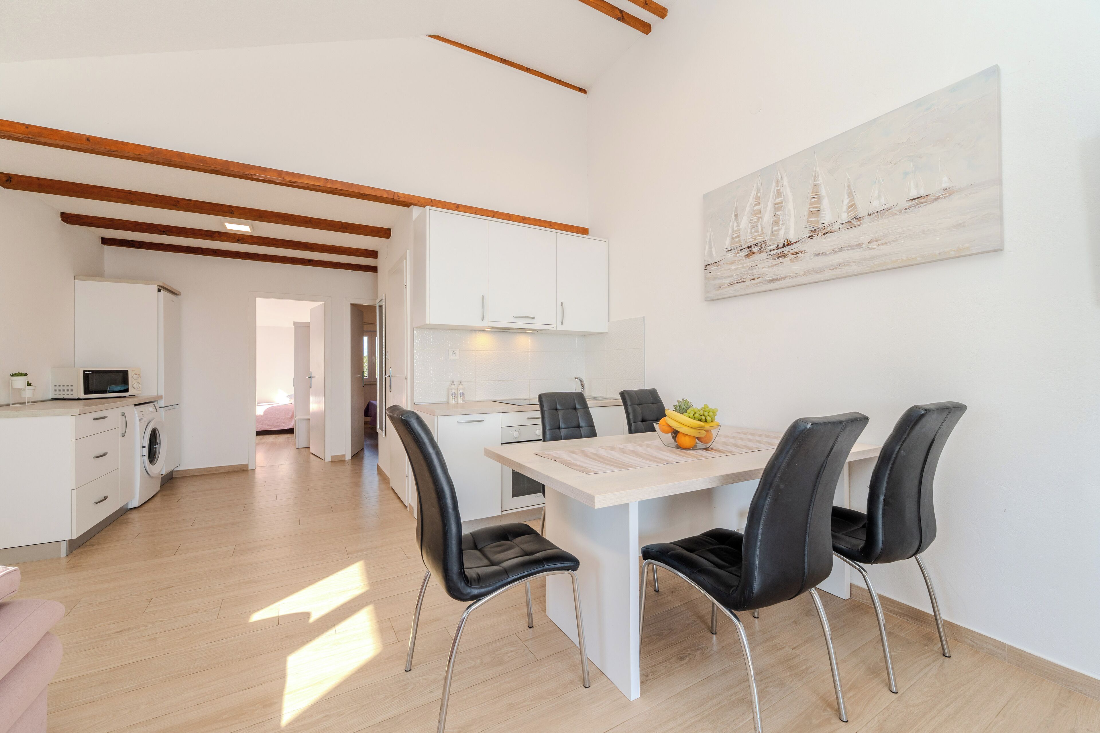 Une cuisine moderne avec des meubles blancs, un coin repas doté d’une table en bois et de chaises noires, et un tableau représentant des voiliers accroché au mur.