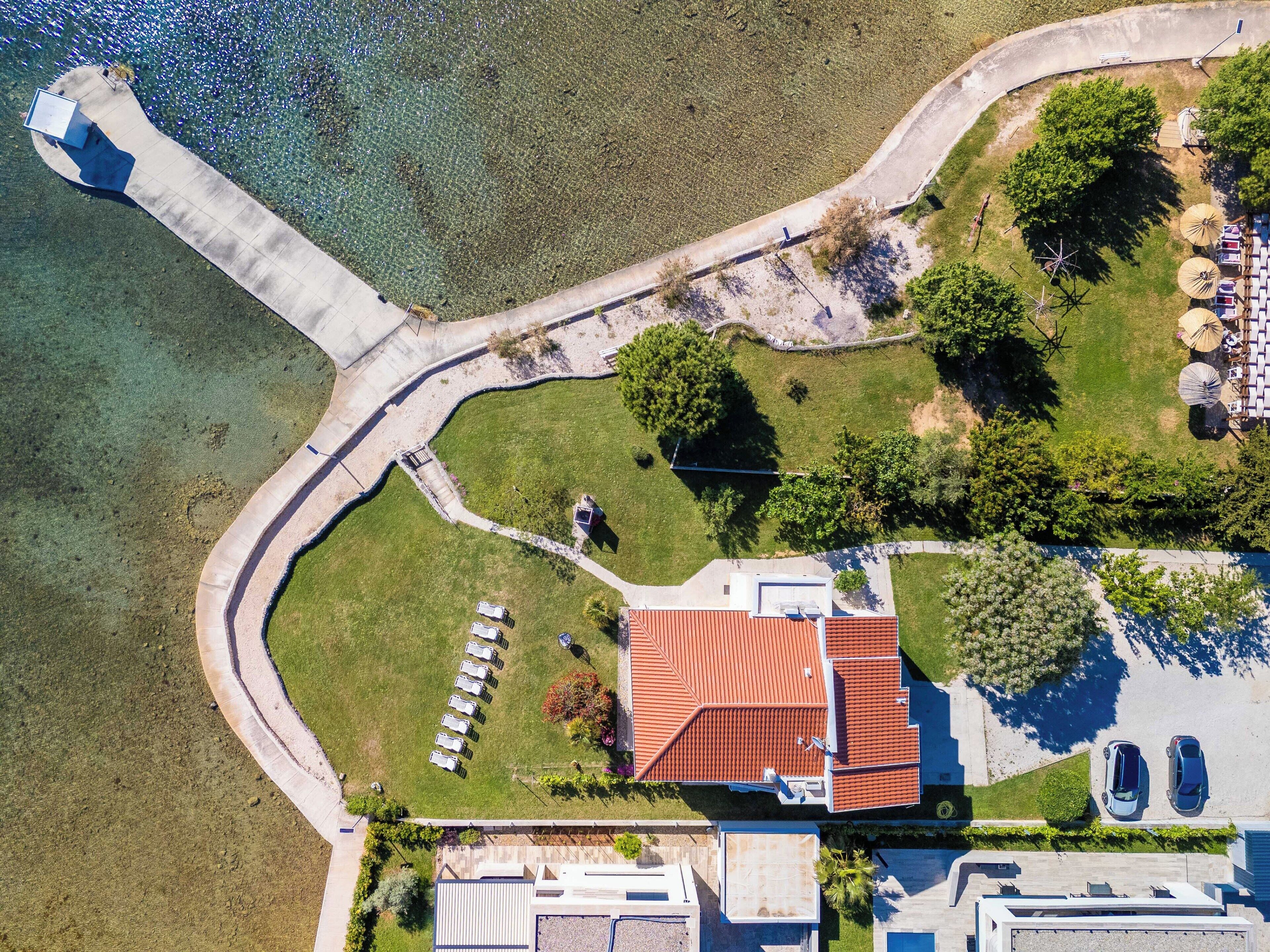 An aerial view of a coastal property with a red-roofed building, a paved walkway, and a parking area.