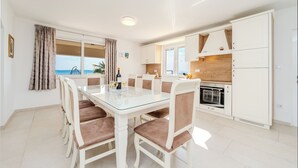 A dining area with a glass table and chairs, a kitchen with white cabinets, and a view of the sea through a window. - Apartments Batela  (Privlaka)