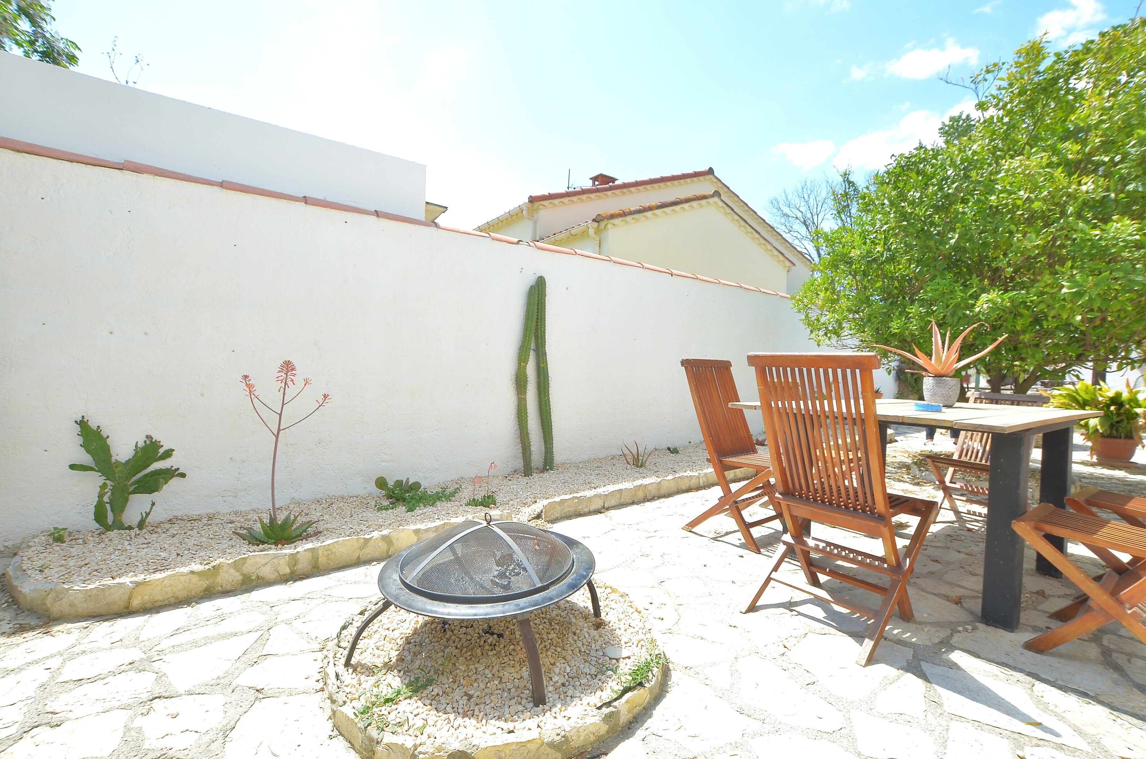 A patio with a table and chairs, a fire pit, and a white wall with a cactus.