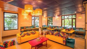 A brightly lit room with yellow sofas, green benches, and tables, featuring large windows and hanging wicker lamps.